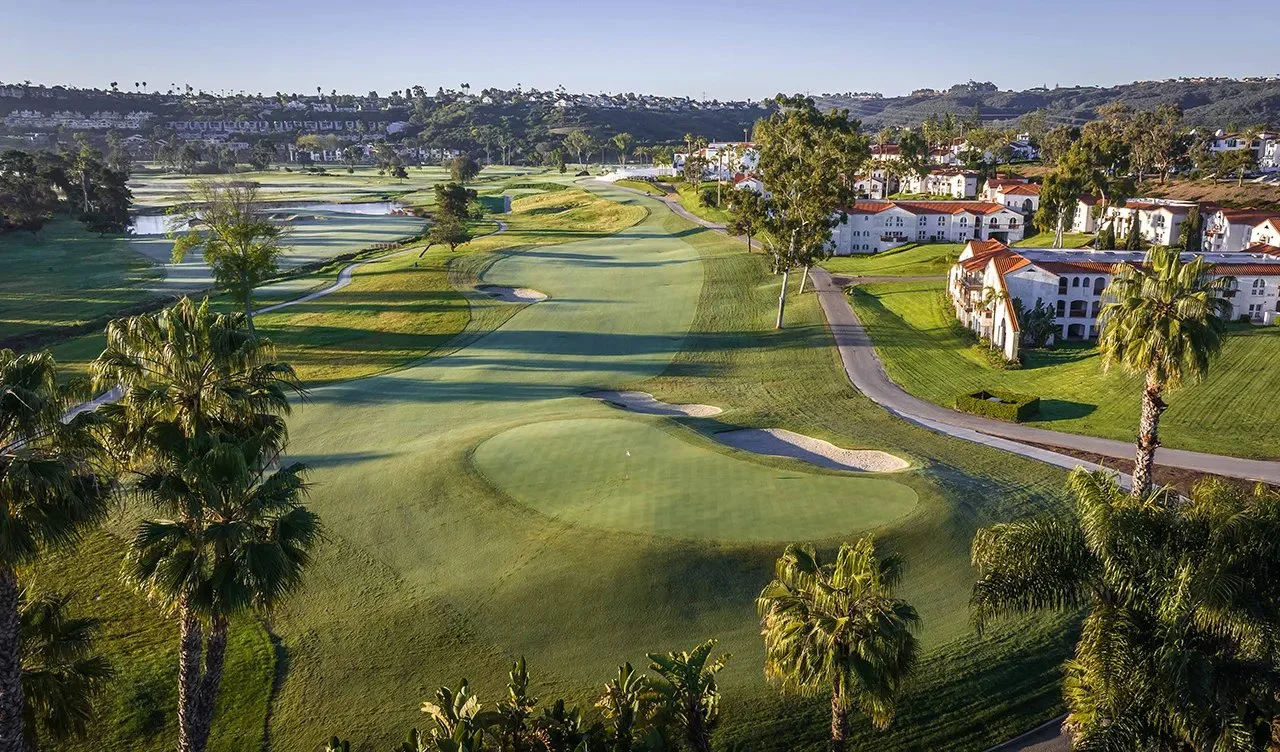 A golf course with green fairways, sand traps, and a putting green, surrounded by palm trees and residential buildings.