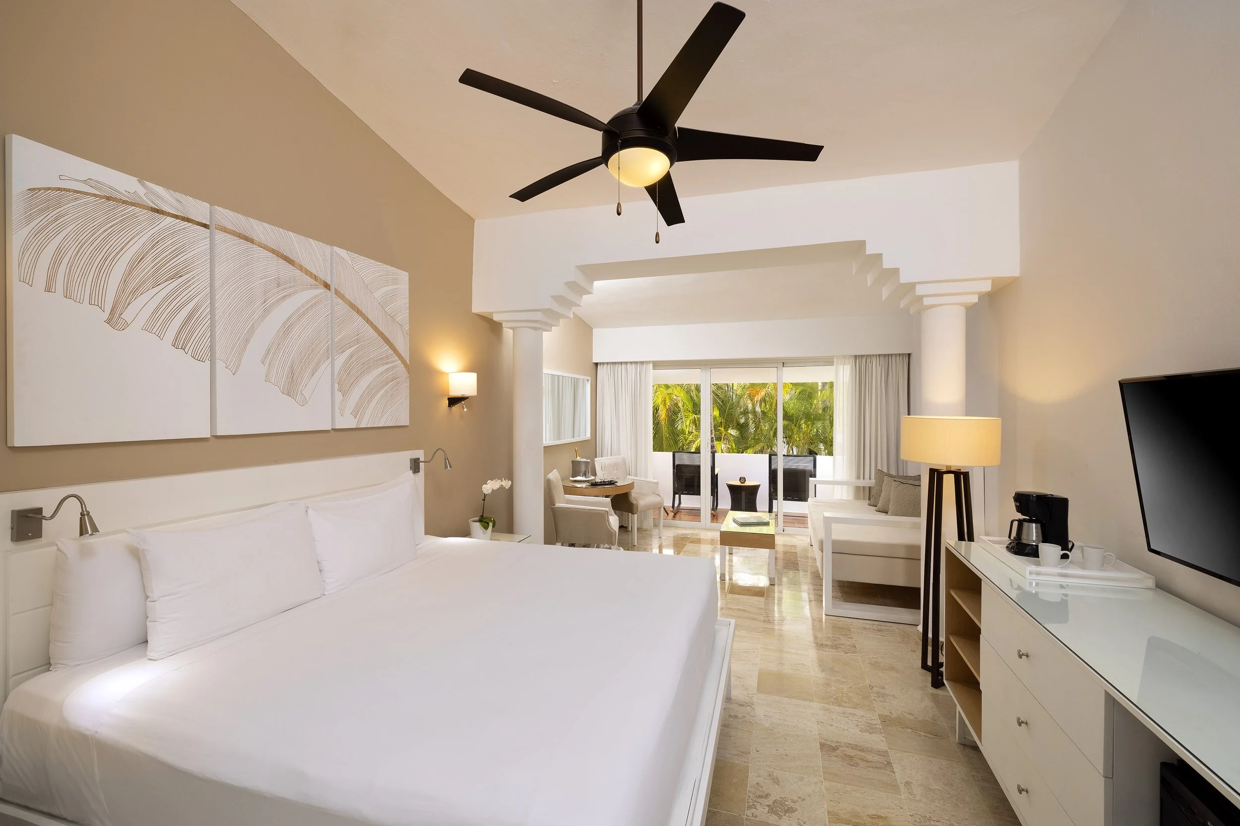 Hotel room with a large bed, wall art, a ceiling fan, a small sitting area near a balcony with tropical greenery, and a dresser with a coffee maker.