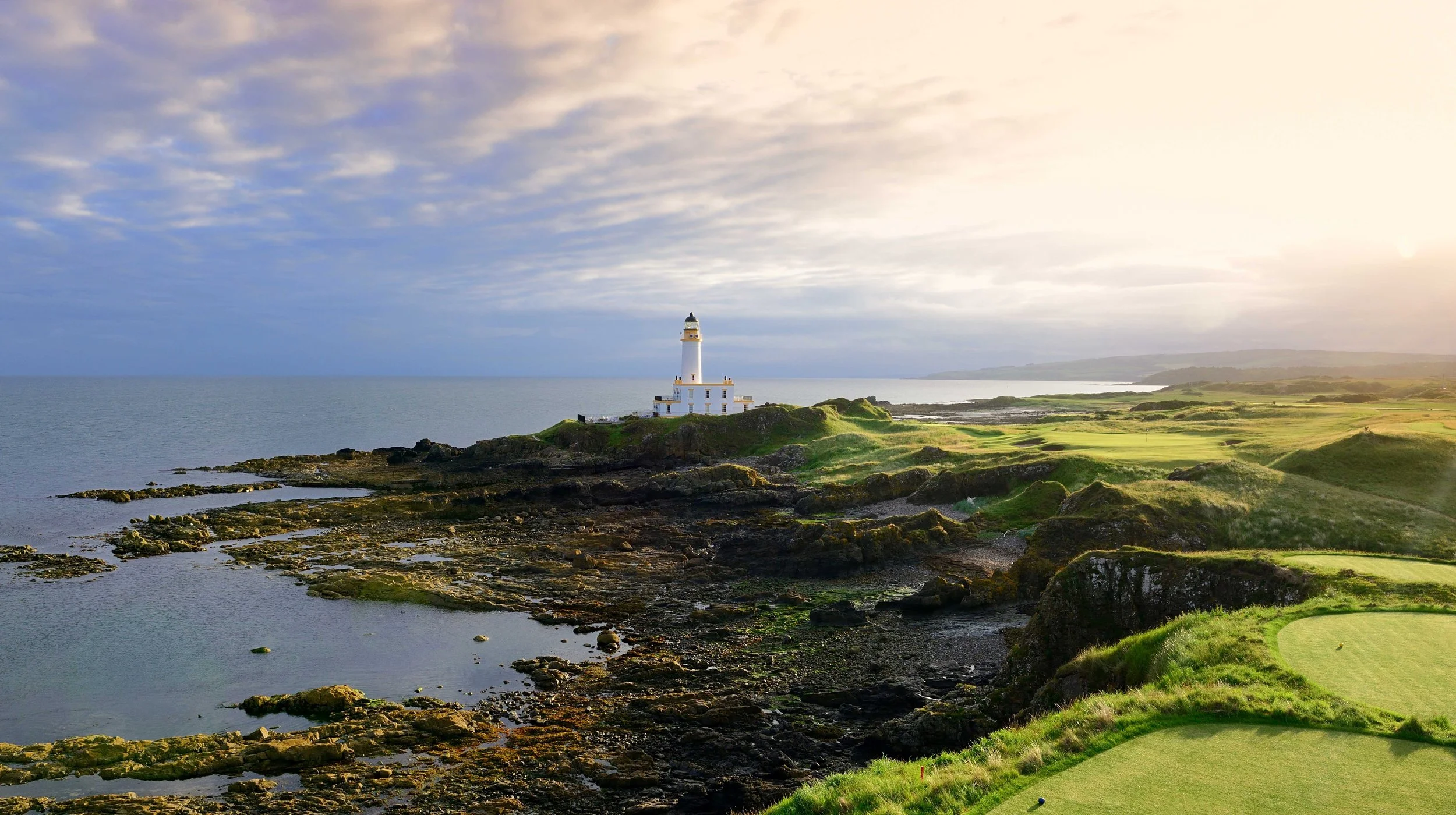 A coastal lighthouse on rocky shoreline with lush green grass and a golf course in the foreground, under a cloudy sky with some sunlight.