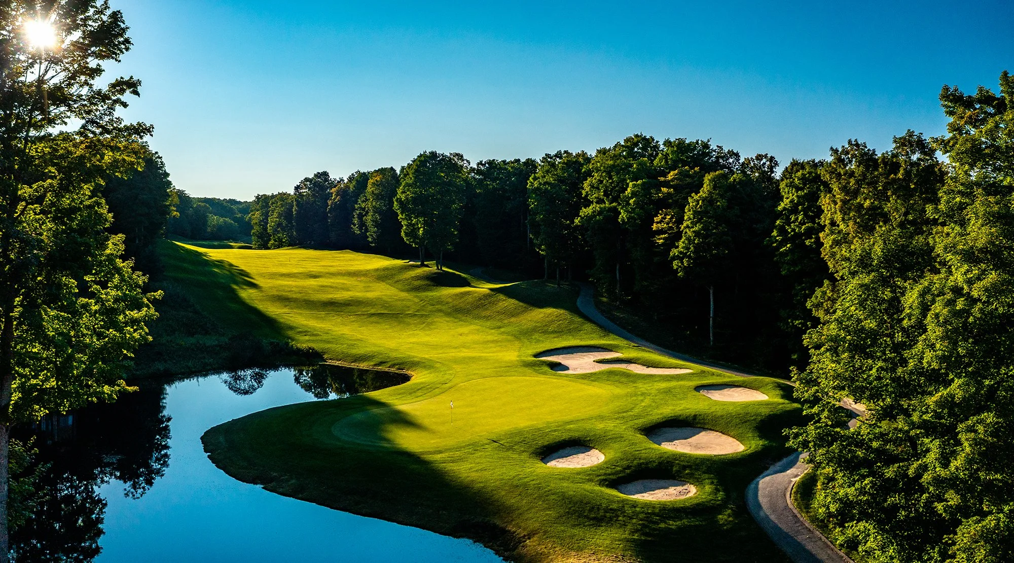 A scenic golf course with a small pond, sand bunkers, and a lush green fairway surrounded by tall trees on a sunny day