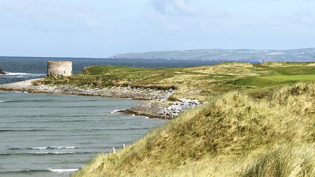Coastal landscape with grassy cliffs, rocky shoreline, and a round stone tower on the coast, with the ocean and distant hills in the background.