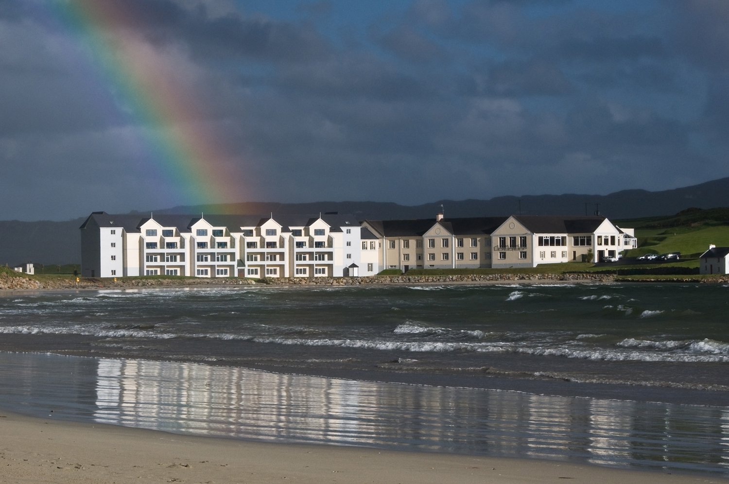 Beach with waves in the foreground, a large modern beachfront apartment building behind, dark clouds overhead, and a rainbow in the sky.