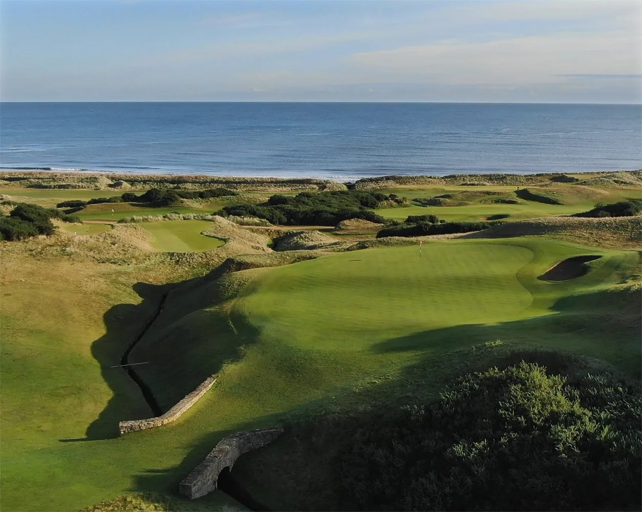 A scenic view of a coastal golf course with green fairways, sand traps, and ocean in the background.