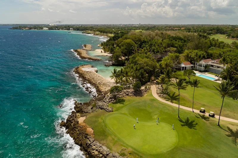 Aerial view of a coastline golf course with palm trees, a green field, and a blue ocean. Three people are visible on the golf green near the shore.
