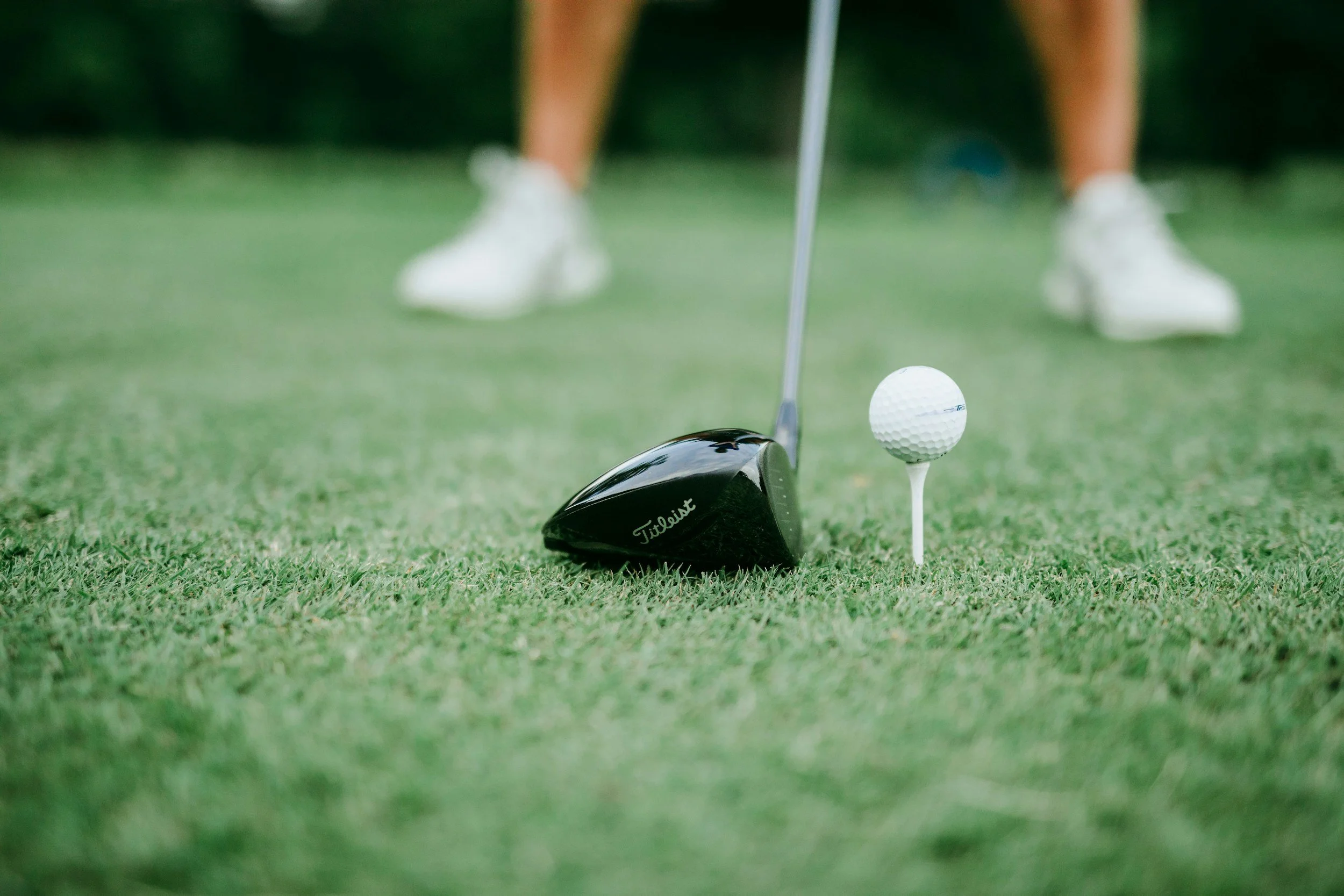 Golf club positioned near a golf ball on a tee on the green, with a person's legs and shoes in the background.