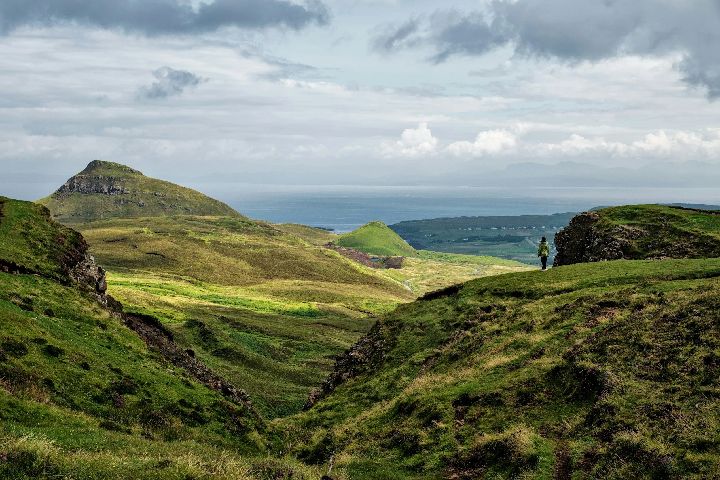 A person walking through lush green rolling hills under cloudy sky, with distant mountains and body of water in the background.