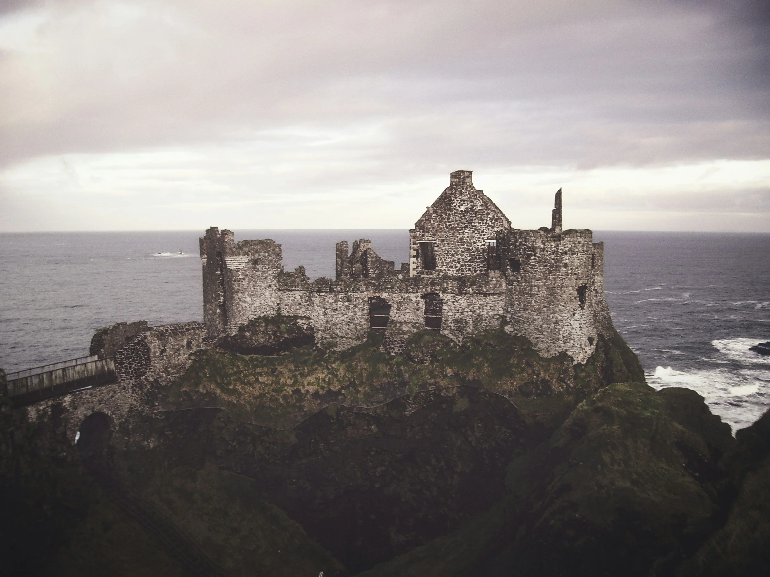 Ruins of a castle on a rocky coastline with the ocean in the background, under a cloudy sky.