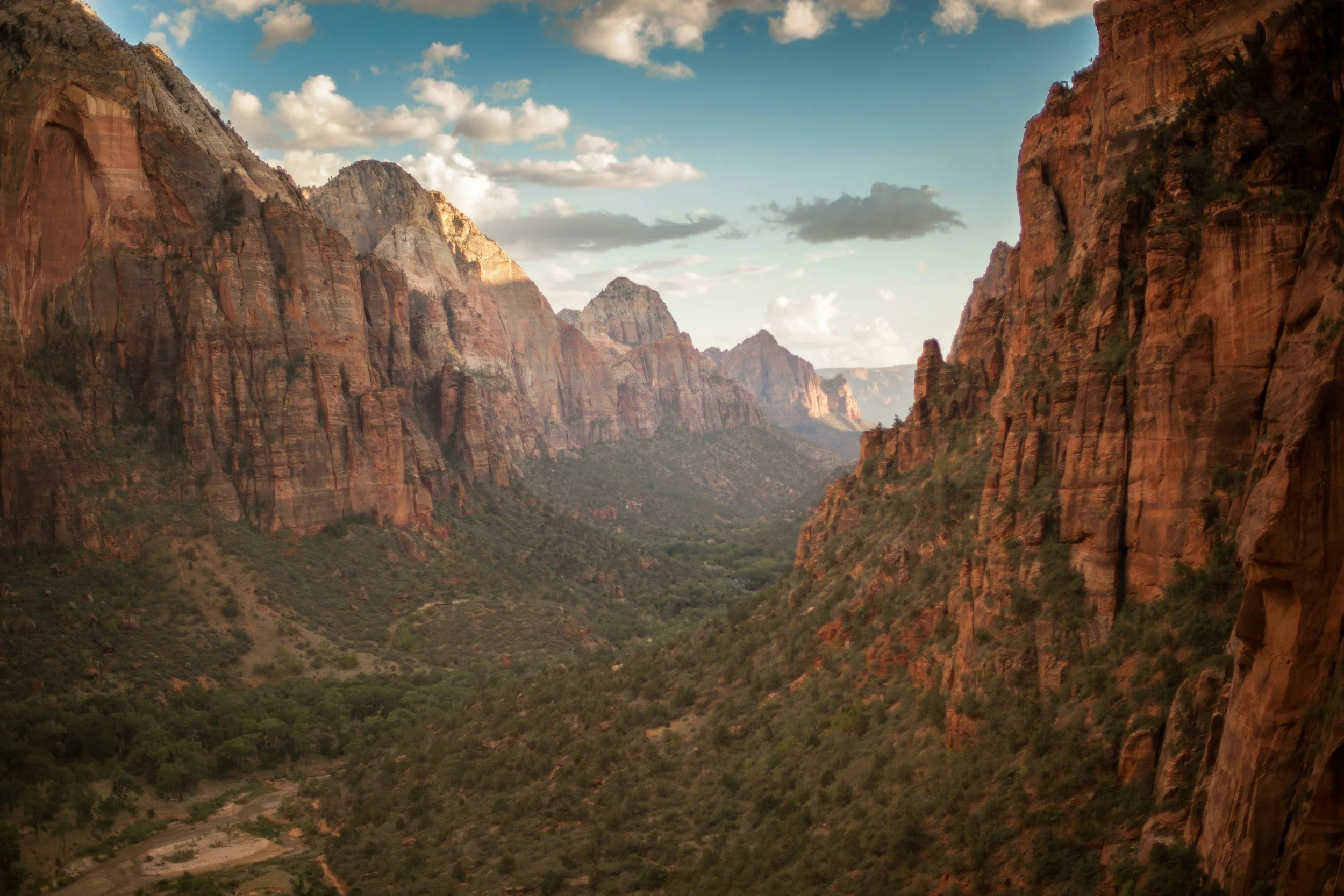 A view of a deep canyon with reddish cliffs on either side and a green valley in the middle, under a partly cloudy sky.