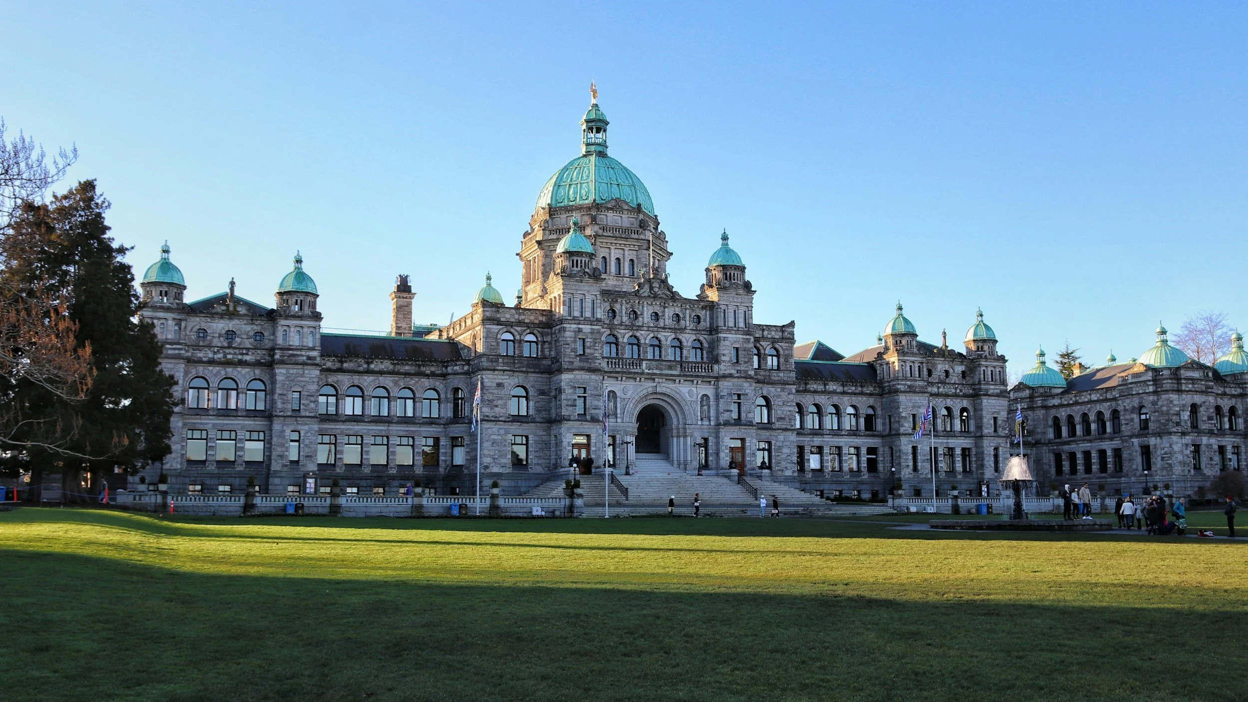 The British Columbia Parliament building in Victoria, British Columbia, Canada, showcasing historic architecture with a large central dome, smaller domes, and a manicured green lawn in the foreground on a clear day.