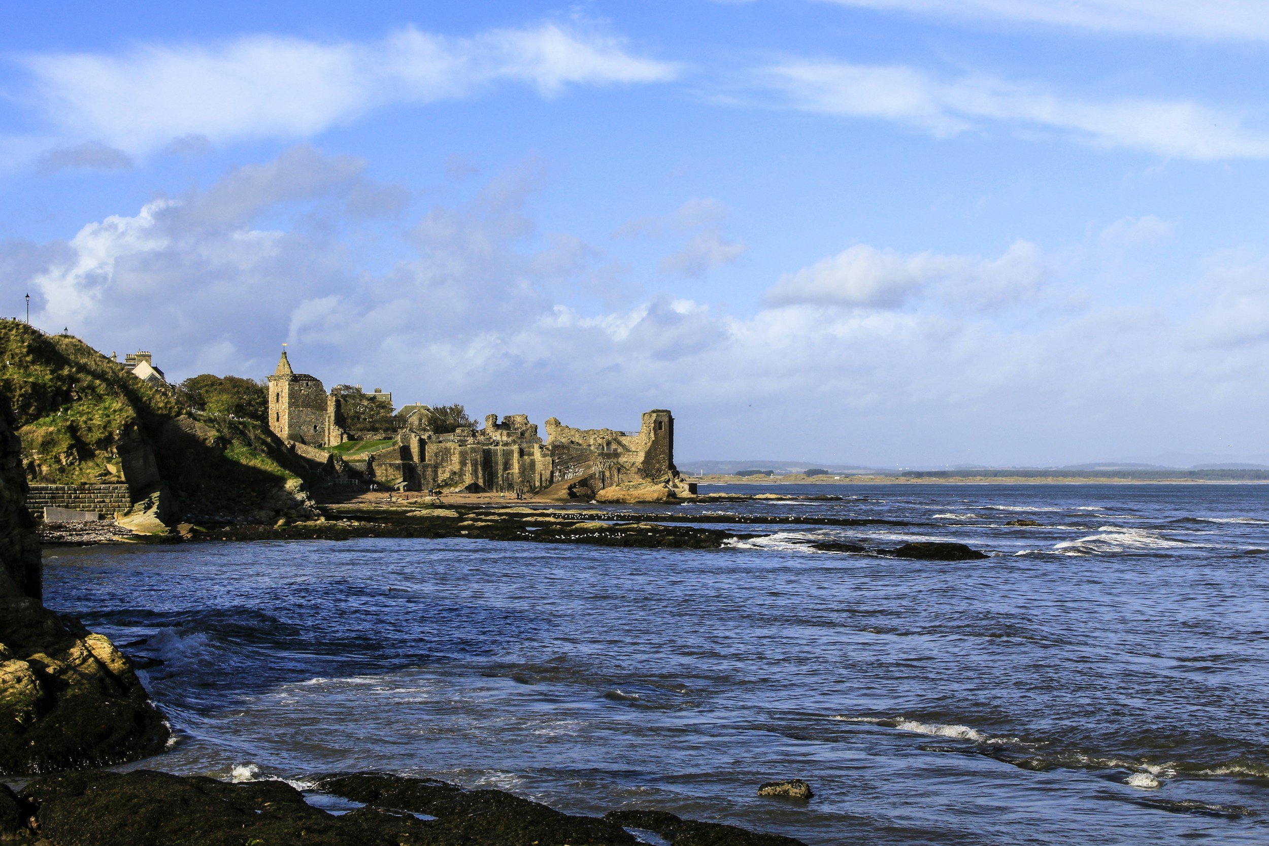 Coastal scene with ruins of an old stone castle and buildings on a hillside, overlooking the ocean with small waves and a partly cloudy sky.