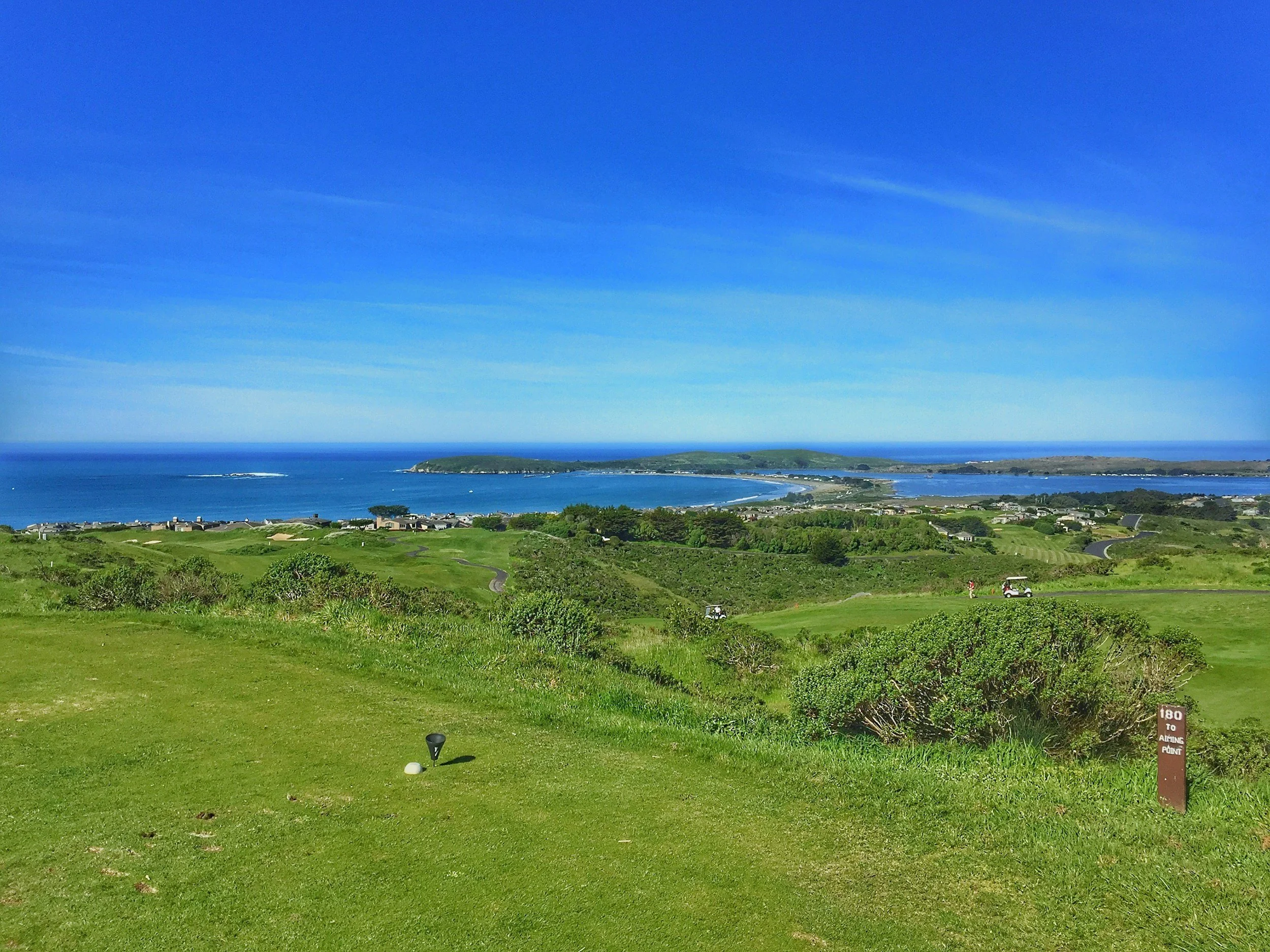 View of a golf course with green fairways and bushes, overlooking a coast with blue ocean, sandy beach, and small islands under a bright blue sky.
