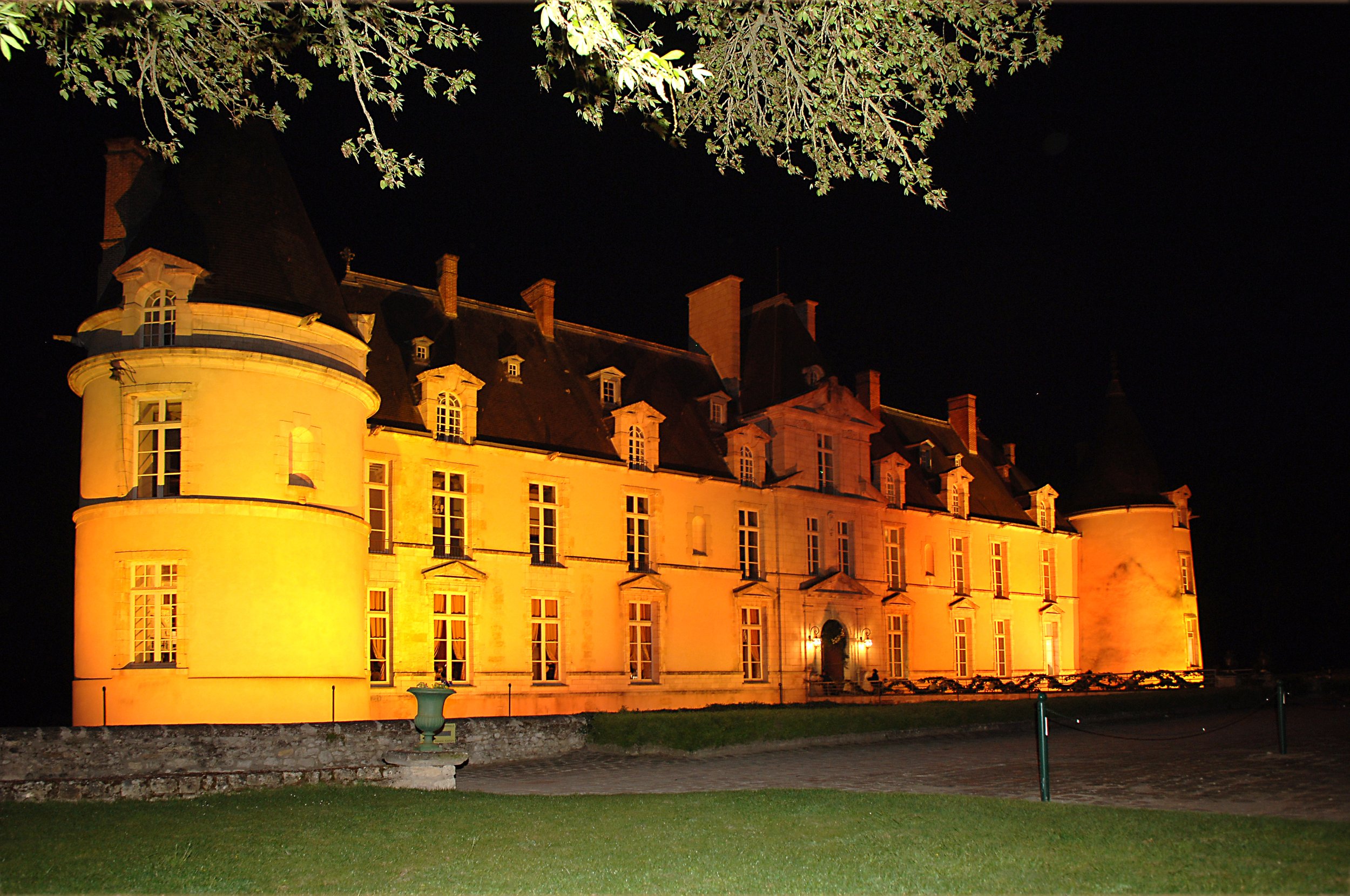A large historic castle at night, illuminated with warm yellow lighting, surrounded by a grass lawn and some trees at the top of the image.