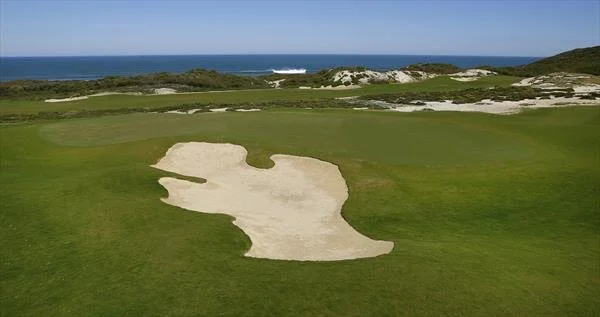A golf course with a sand trap, green grass, near the coast with ocean and dunes in the background.