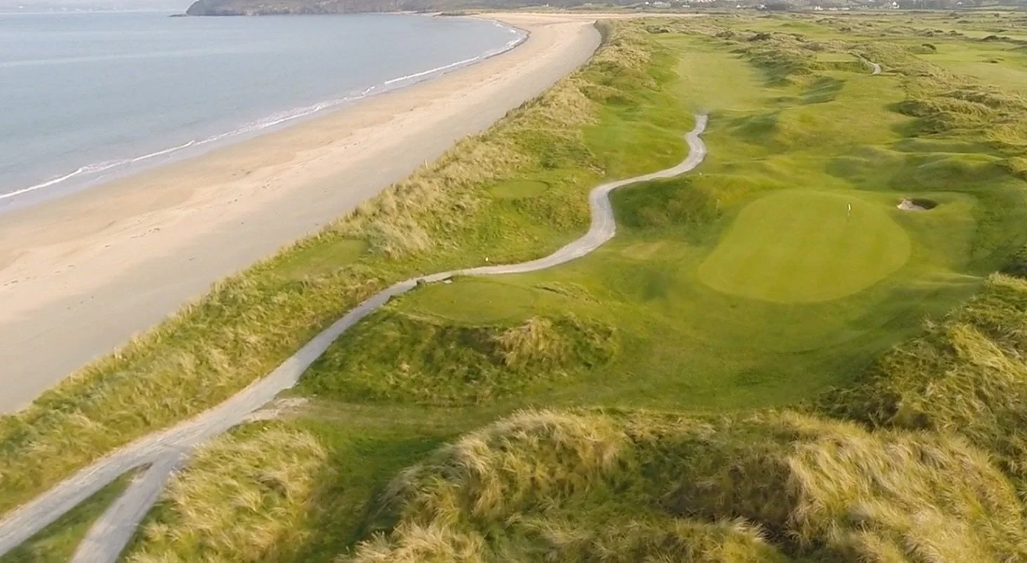 Aerial view of a golf course along a coastline with a sandy beach and ocean on the left side, featuring green fairways and paths.