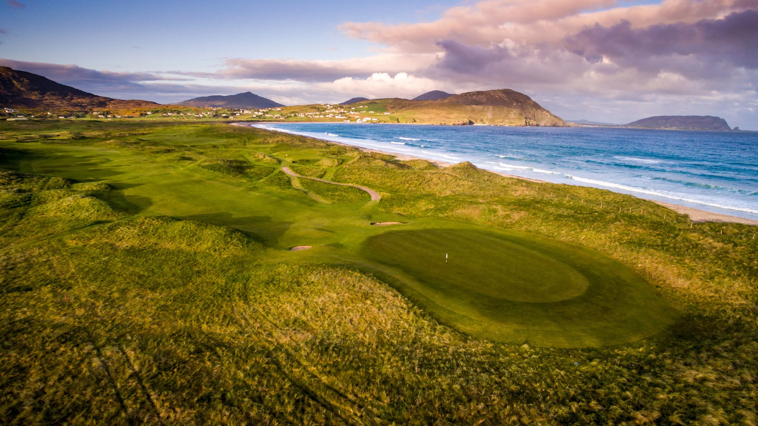 A scenic coastal landscape featuring a golf course with green fairways and sand traps, overlooking the ocean with waves, and rolling hills in the background under a partly cloudy sky.