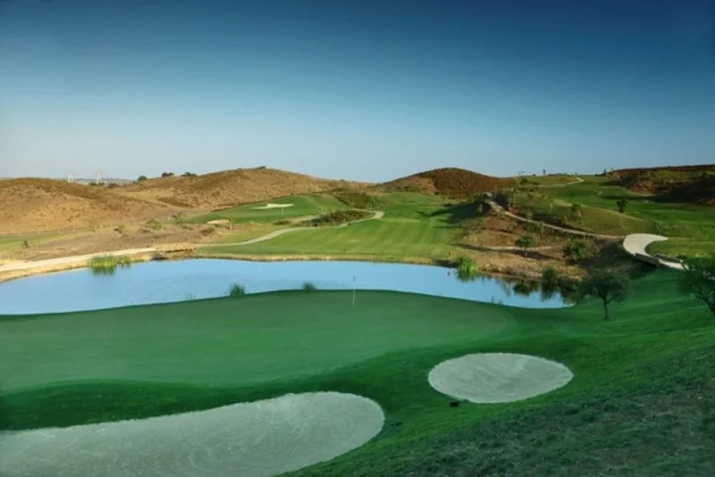 A golf course with green fairways, sand bunkers, a pond, and surrounding hills under a clear blue sky.