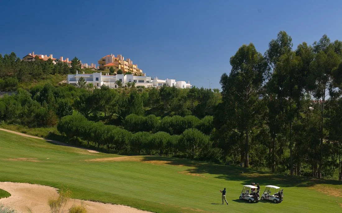 Golf course with two golf carts and a person playing golf under a clear blue sky, with green trees and buildings on a hillside in the background.
