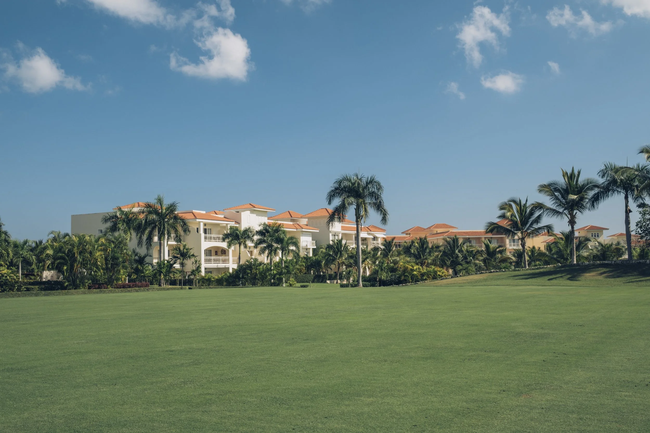 A view of a luxurious resort with white buildings and red-tiled roofs, surrounded by palm trees, green lawns, and a clear blue sky with a few clouds.