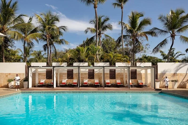Swimming pool with sunny lounge chairs and cabanas, palm trees in the background, and clear blue sky.