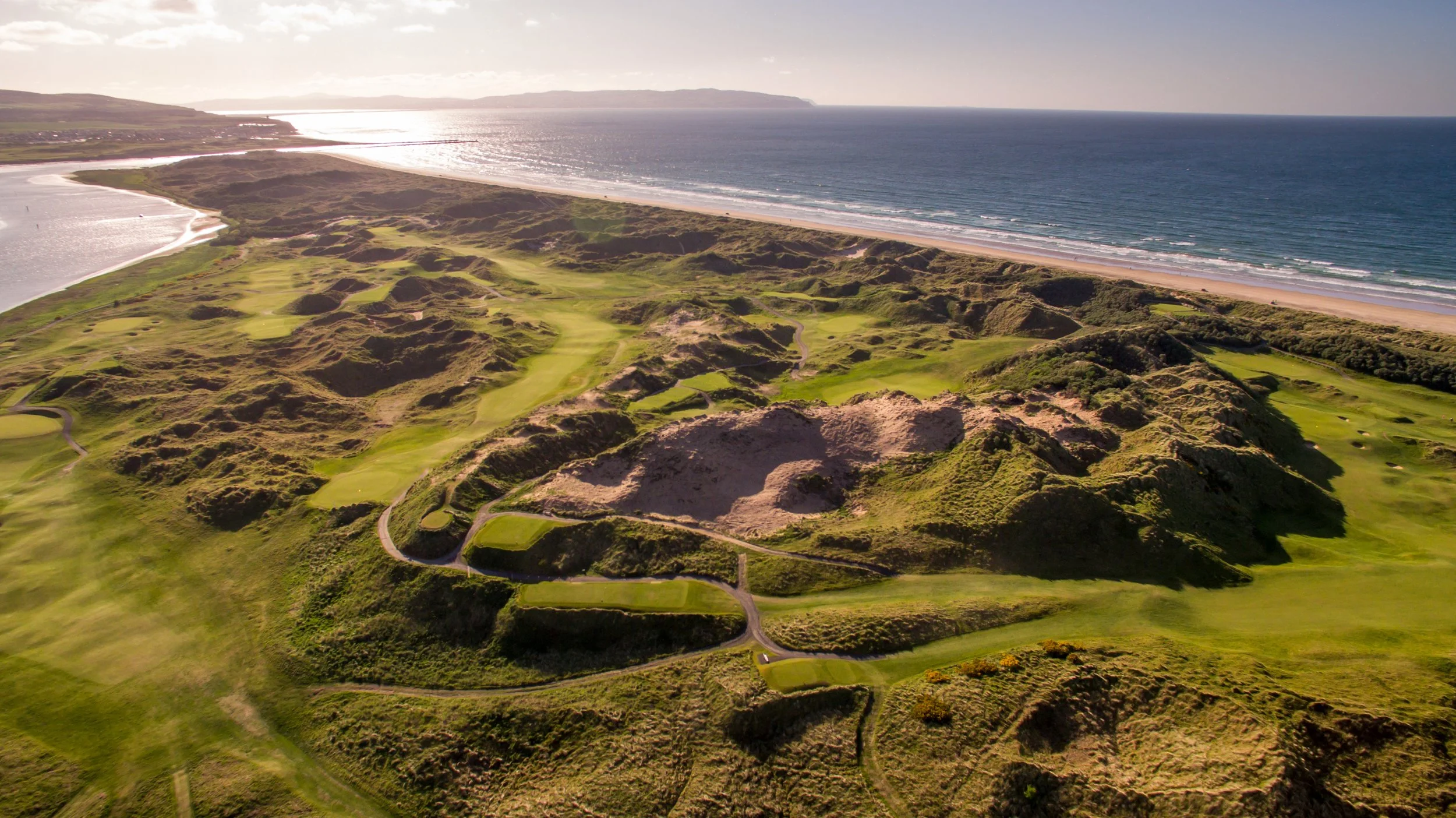 Aerial view of a coastal golf course with rolling hills, green fairways, sand dunes, and a nearby beach and ocean in the background.