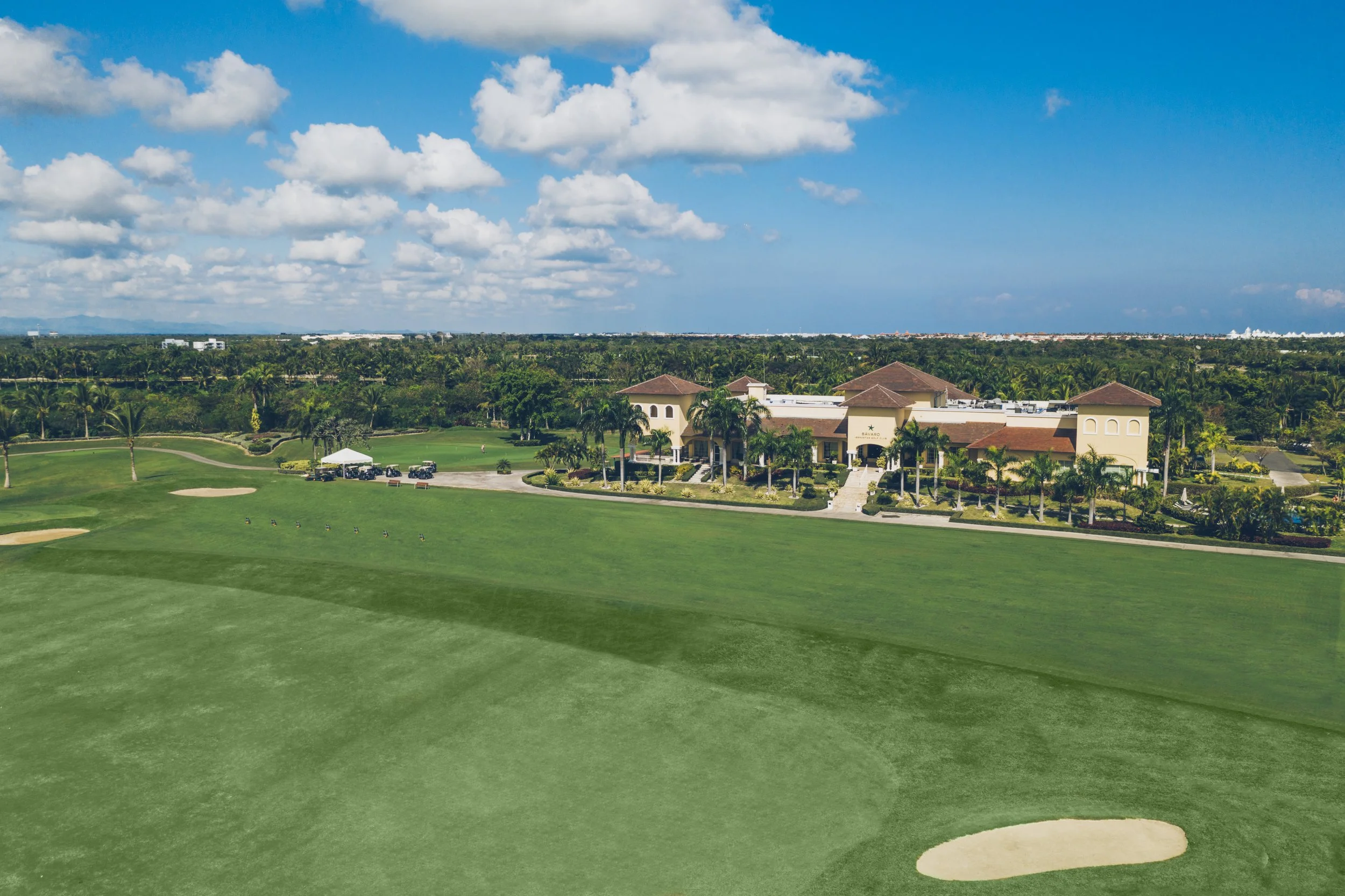 Aerial view of a golf course with green fairways, sand traps, and a clubhouse surrounded by palm trees under a blue sky with scattered clouds.