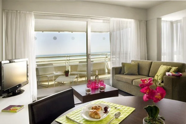 Living room with a large window view of the ocean, a beige sofa, a coffee table with bottles and glasses, a dining table with a plate of croissants and a pink flower arrangement.
