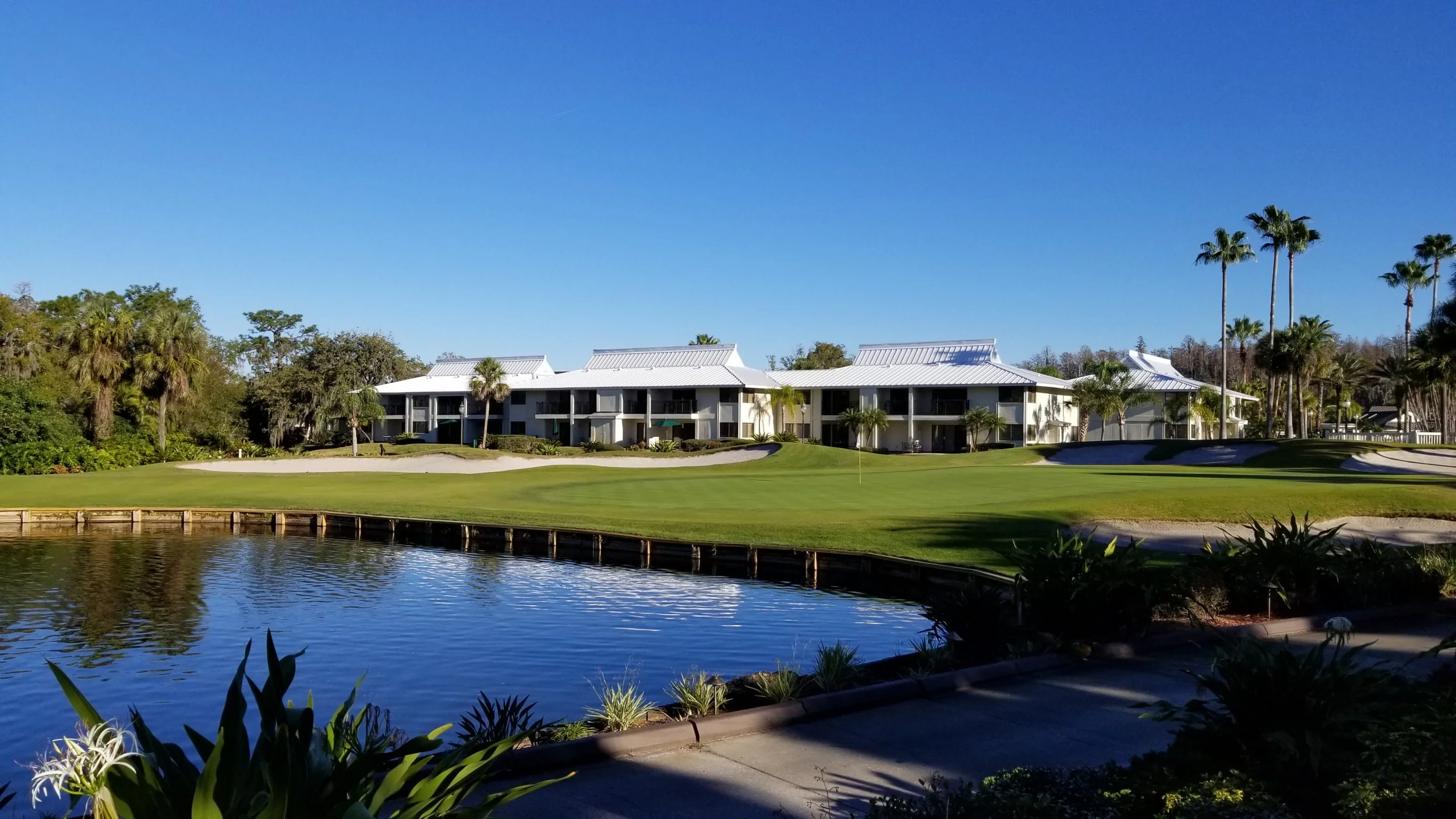A golf course with a water hazard, sand traps, and a multi-unit building with pitched roofs and balconies, surrounded by trees and palm trees, under a clear blue sky.
