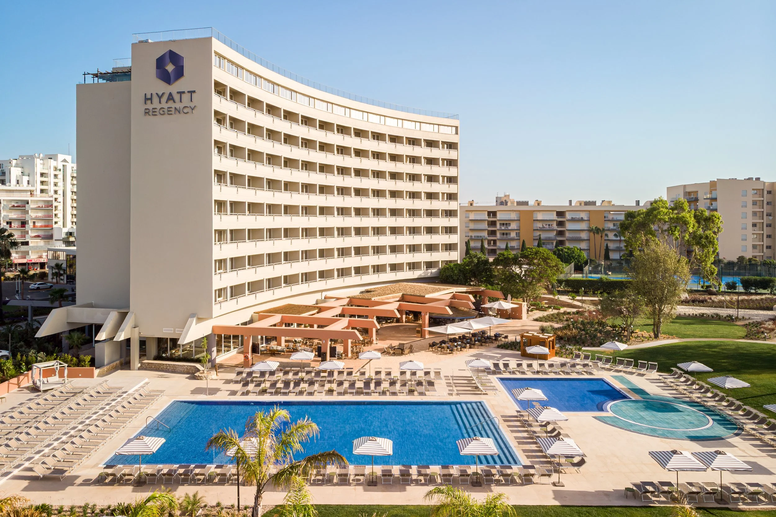 View of the Hyatt Regency hotel with a large swimming pool, poolside lounge chairs with umbrellas, nearby greenery, and surrounding apartment buildings under a clear blue sky.