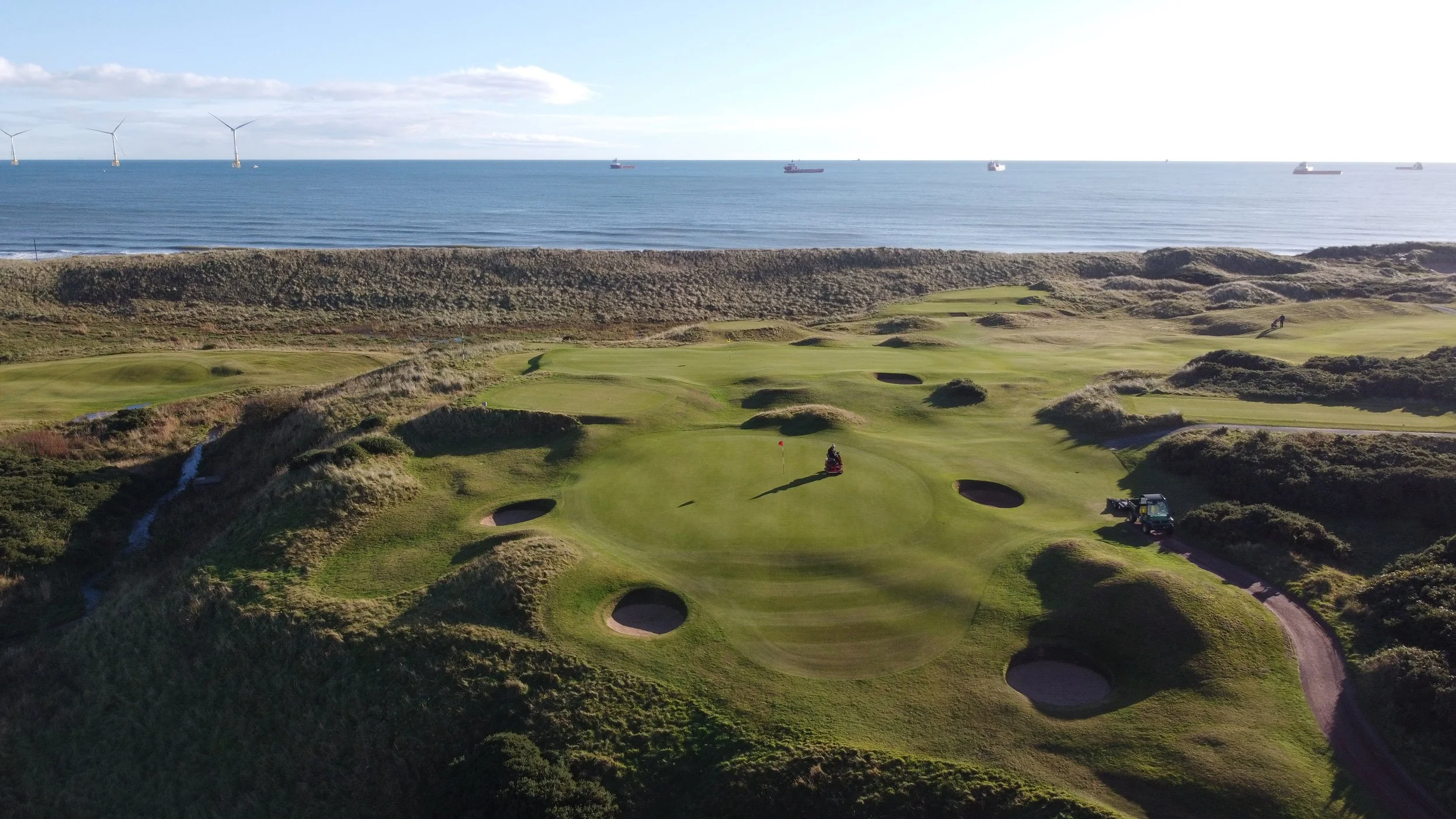Aerial view of a golf course near the beach with wind turbines and ships in the background, showcasing lush green fairways, sand bunkers, and a cart on the course.