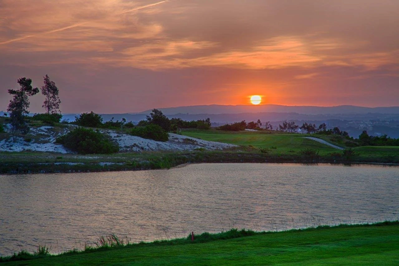 Sunset over a river with grassy banks, trees, and hilly landscape in the background.