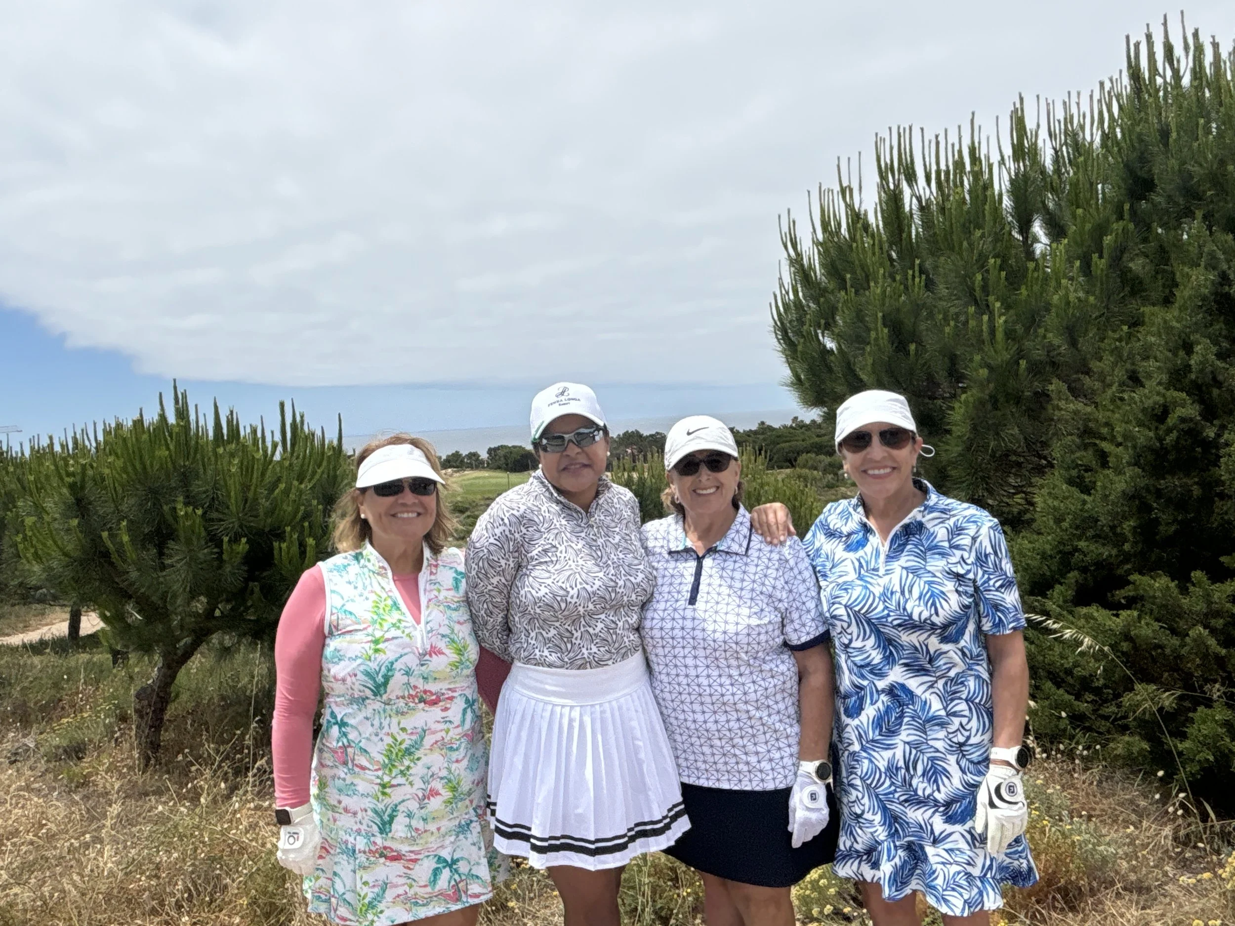 Four women in golf attire standing outdoors, smiling, wearing hats and sunglasses, with trees and a cloudy sky in the background.