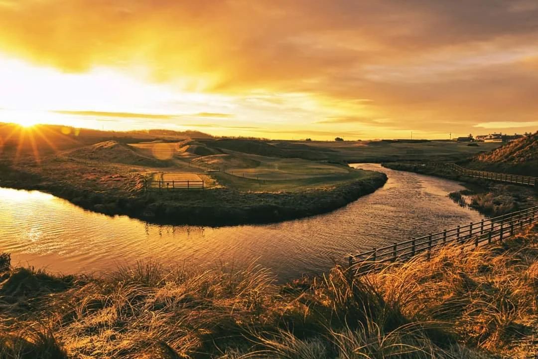 Sunset over a golf course with a winding waterway, grassy hills, wooden fences, and a cloudy sky.