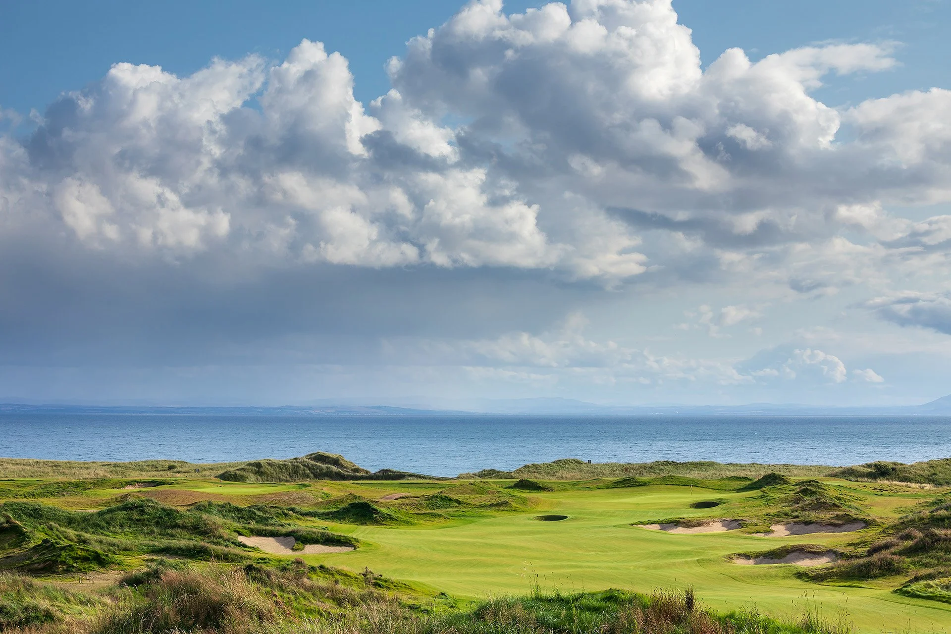 Scenic view of a golf course along the coast with lush green fairways, sand bunkers, and rolling terrain under a partly cloudy sky.