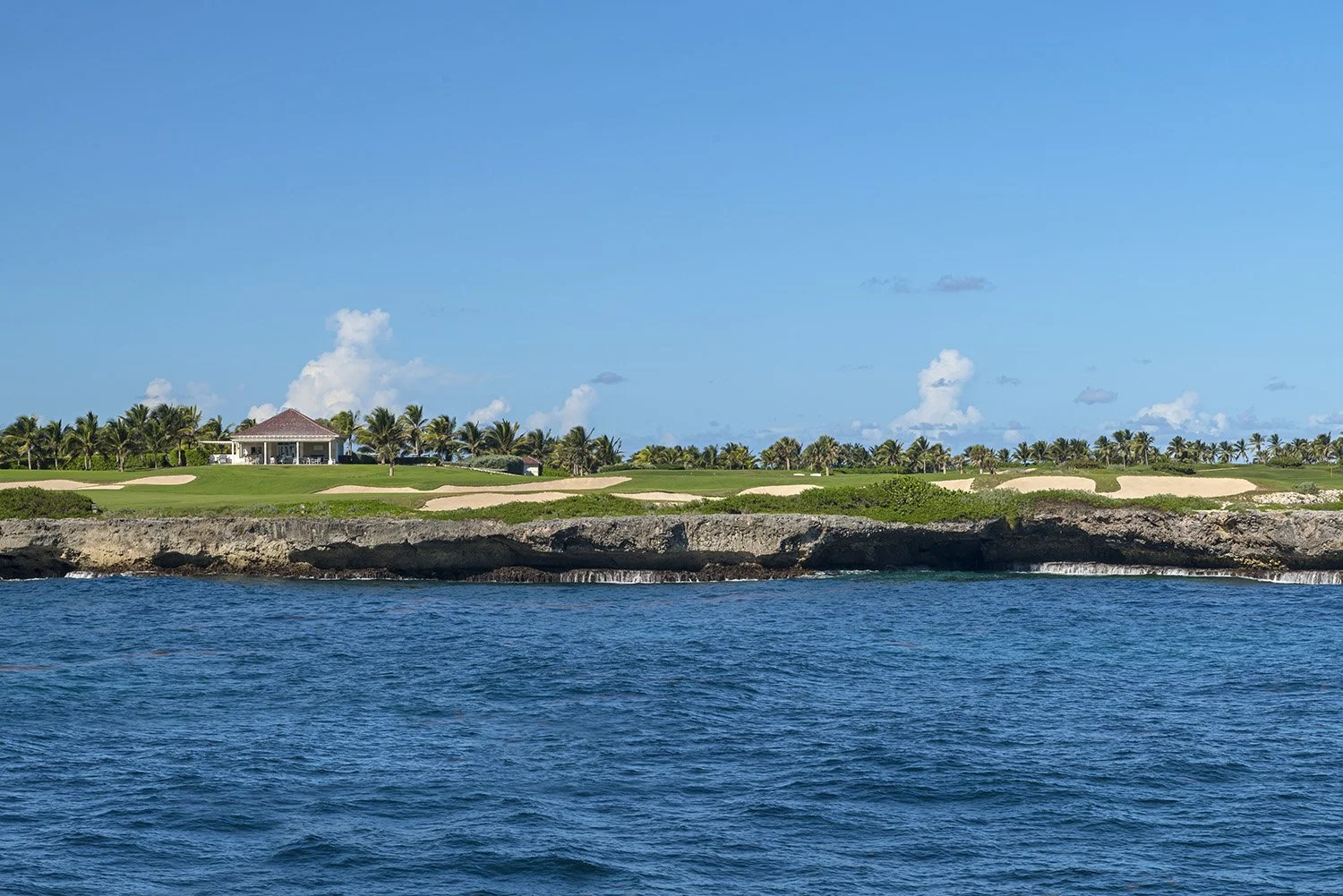 Coastal landscape with a golf course, palm trees, and a house on a rocky shoreline under a clear blue sky.