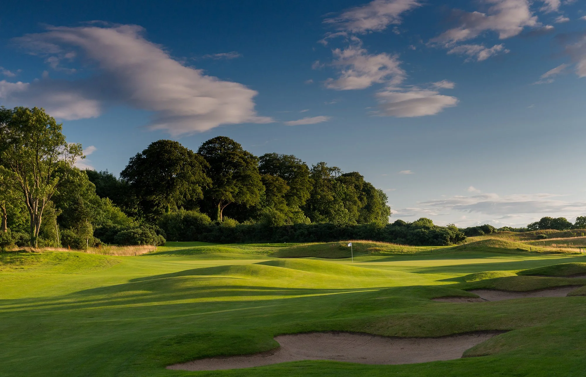 A golf course with green fairways, sand traps, and a flag on the putting green, surrounded by trees under a partly cloudy sky.