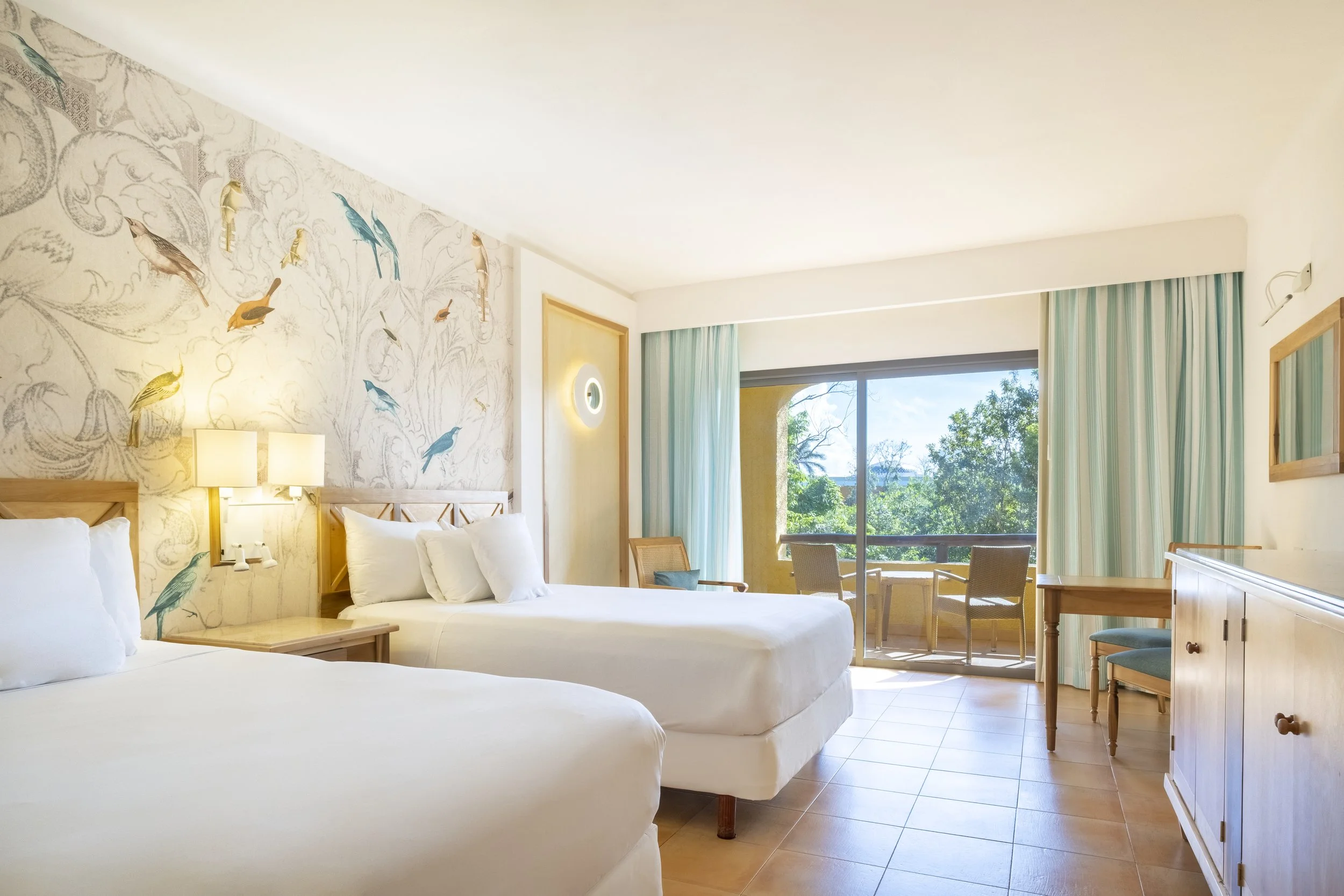 A bright hotel room with two white beds, a wall decorated with bird illustrations, a sliding glass door leading to a balcony with outdoor chairs, and a view of green trees outside.
