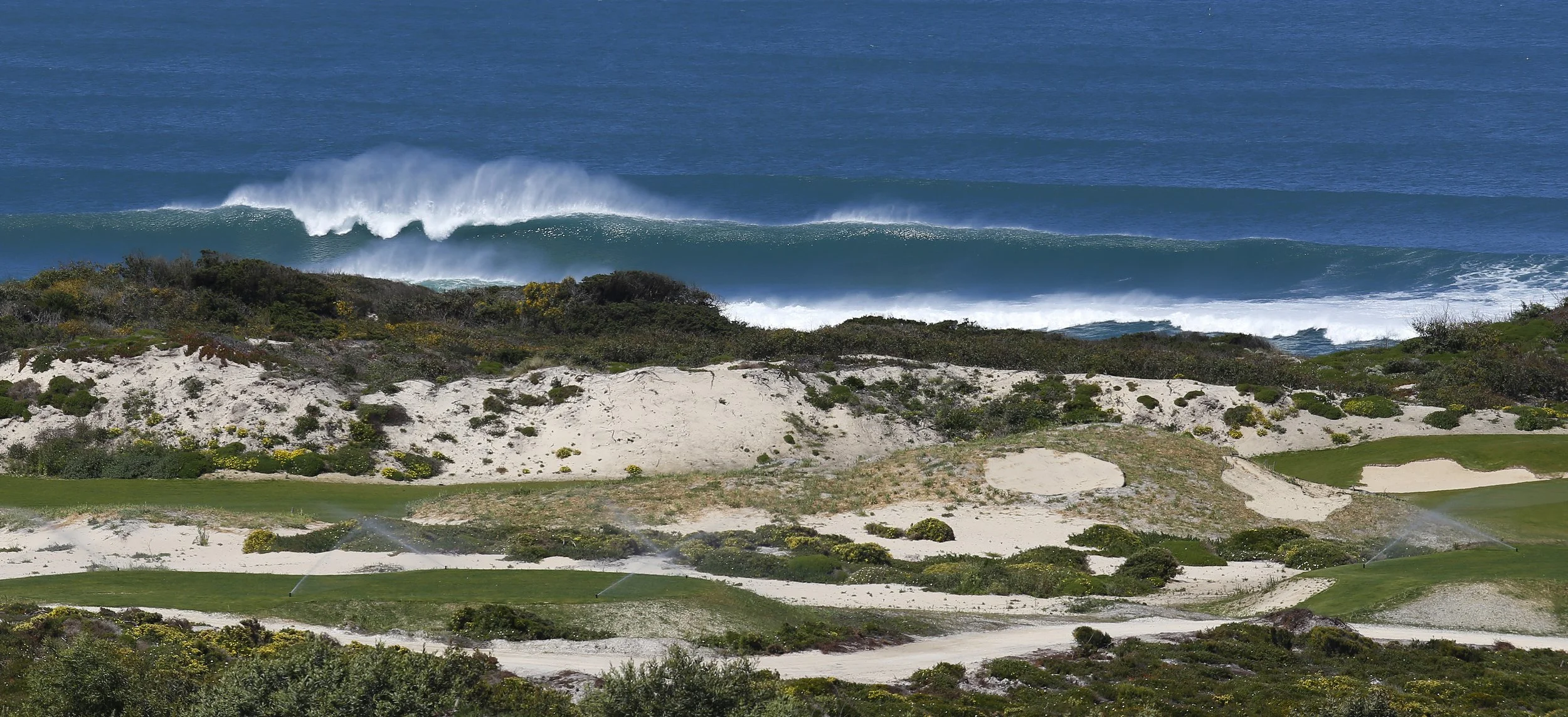 A scenic view of the ocean with large waves crashing near the shore, seen from a coastal area with sandy dunes, green vegetation, and a golf course with sprinklers.