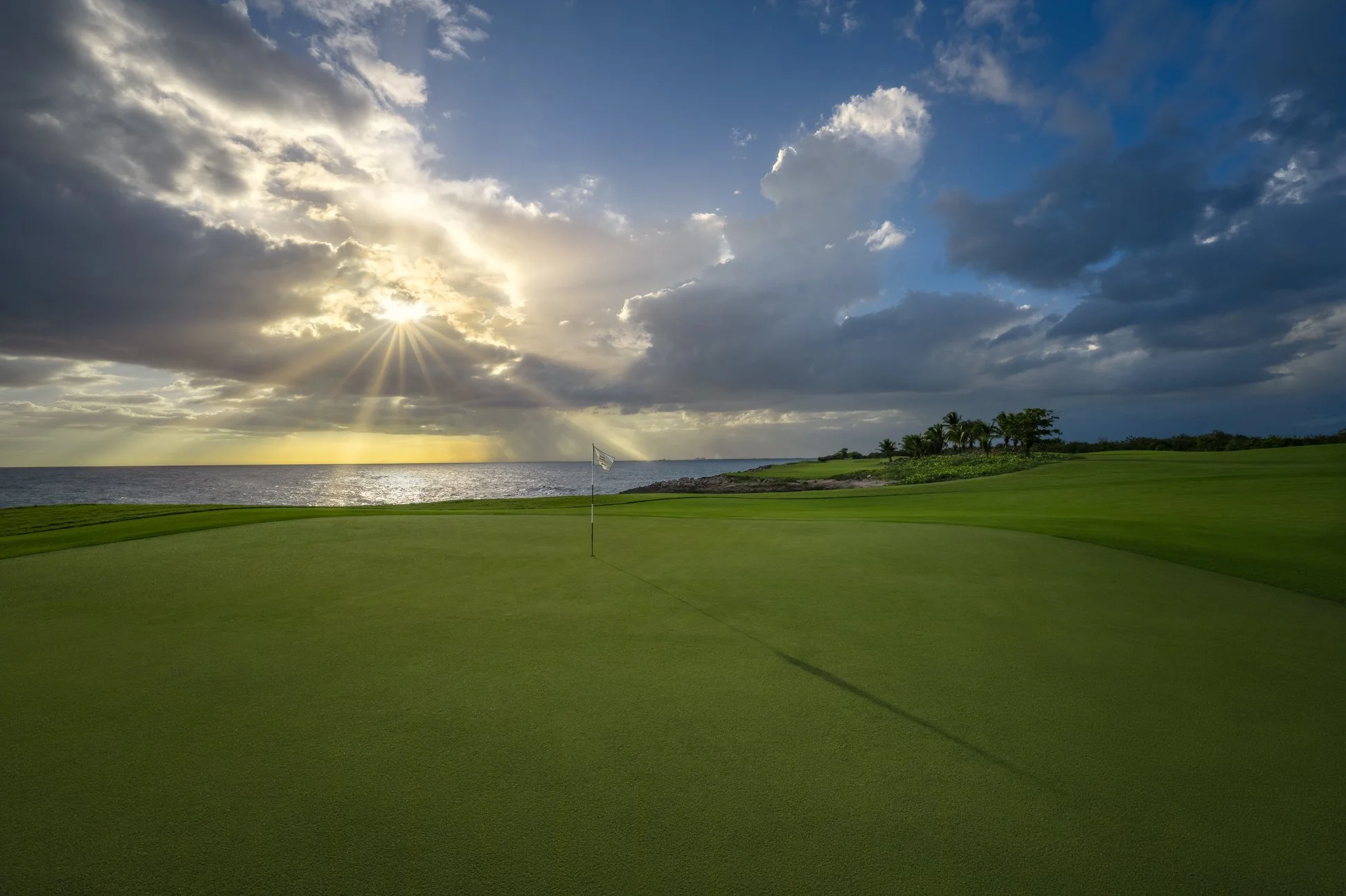 A golf course near the ocean at sunset with a flag on the green, clouds in the sky, and sunlight breaking through.