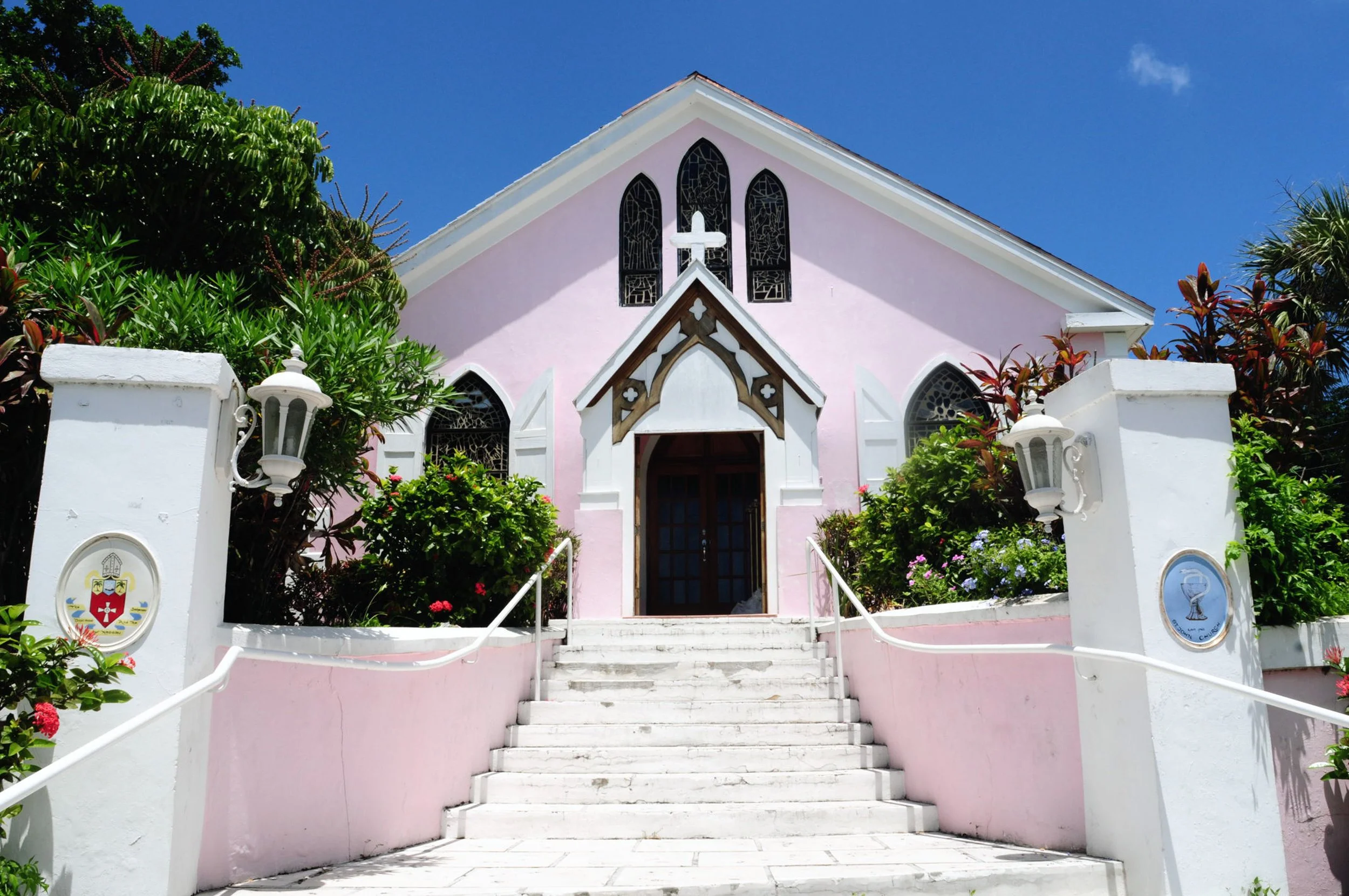 A pink church with white accents, stained glass windows, a cross on the front gable, and a staircase leading up to the entrance, surrounded by green and flowering plants under a blue sky.