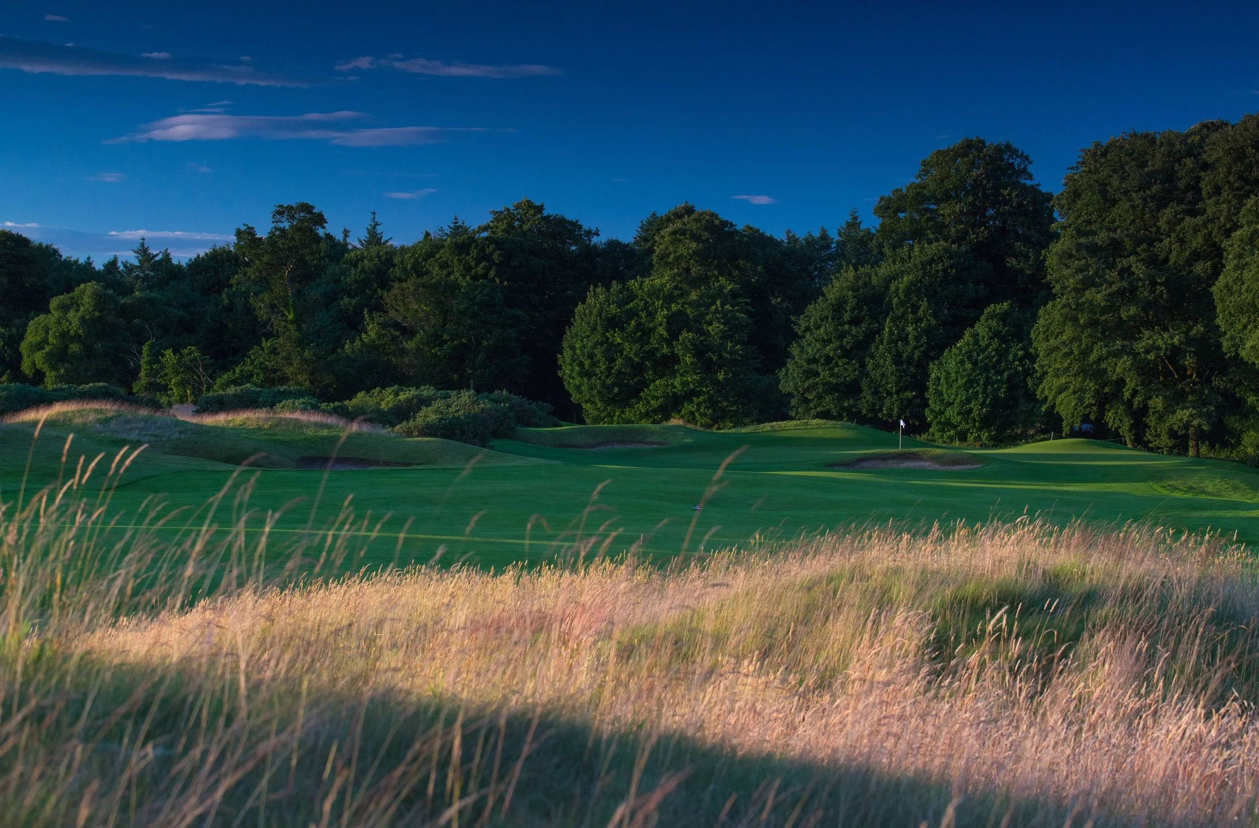 A golf course with green grass, sand bunkers, and a flag on the putting green, surrounded by trees under a blue sky with clouds.