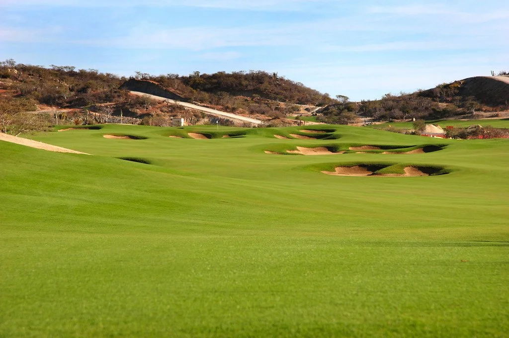 A golf course with green fairways and several sand bunkers, surrounded by hilly terrain and sparse trees under a blue sky.