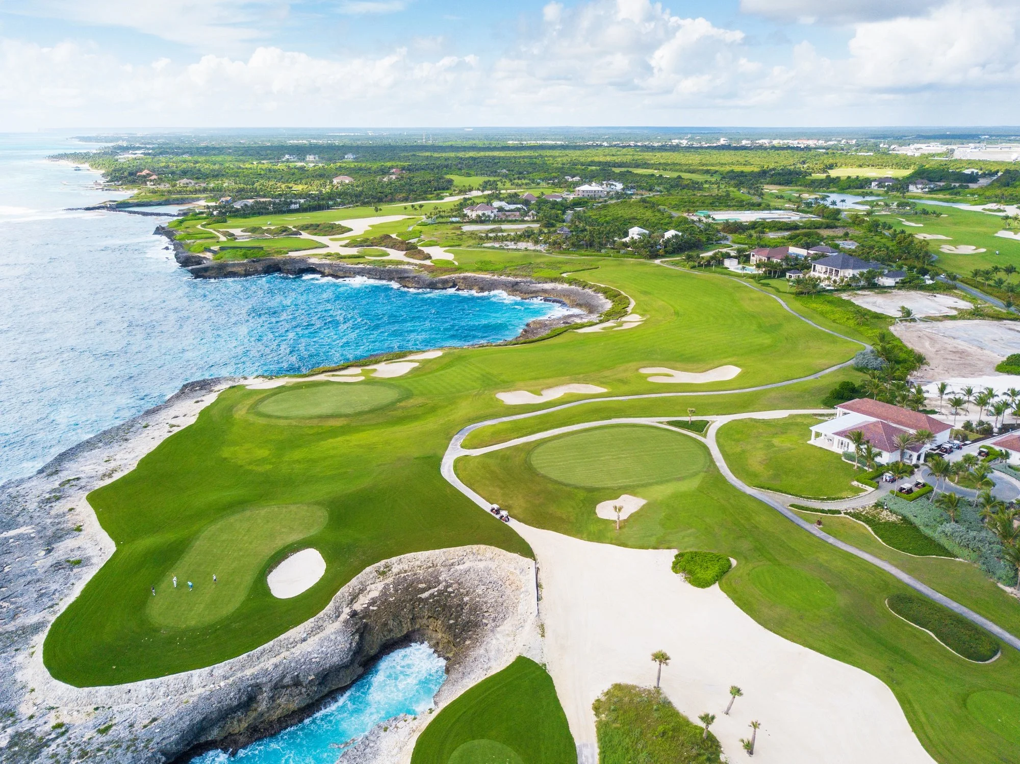 Aerial view of a coastal golf course with lush green fairways, sand bunkers, and rocky shoreline, overlooking the ocean on a partly cloudy day.