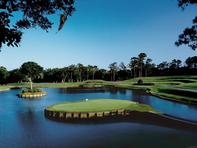 A golf course with water hazards, surrounded by trees, under a clear blue sky.