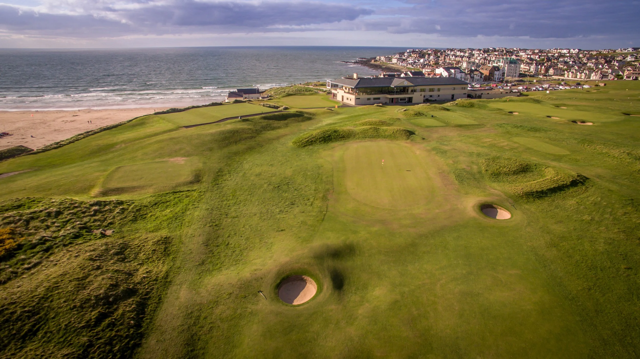 Aerial view of a coastal golf course with sand traps and green fairways, near the beach and ocean, with a residential area in the background and cloudy sky overhead.