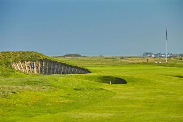 A golf course with a green, sand bunker, and flagstick, under a clear blue sky.