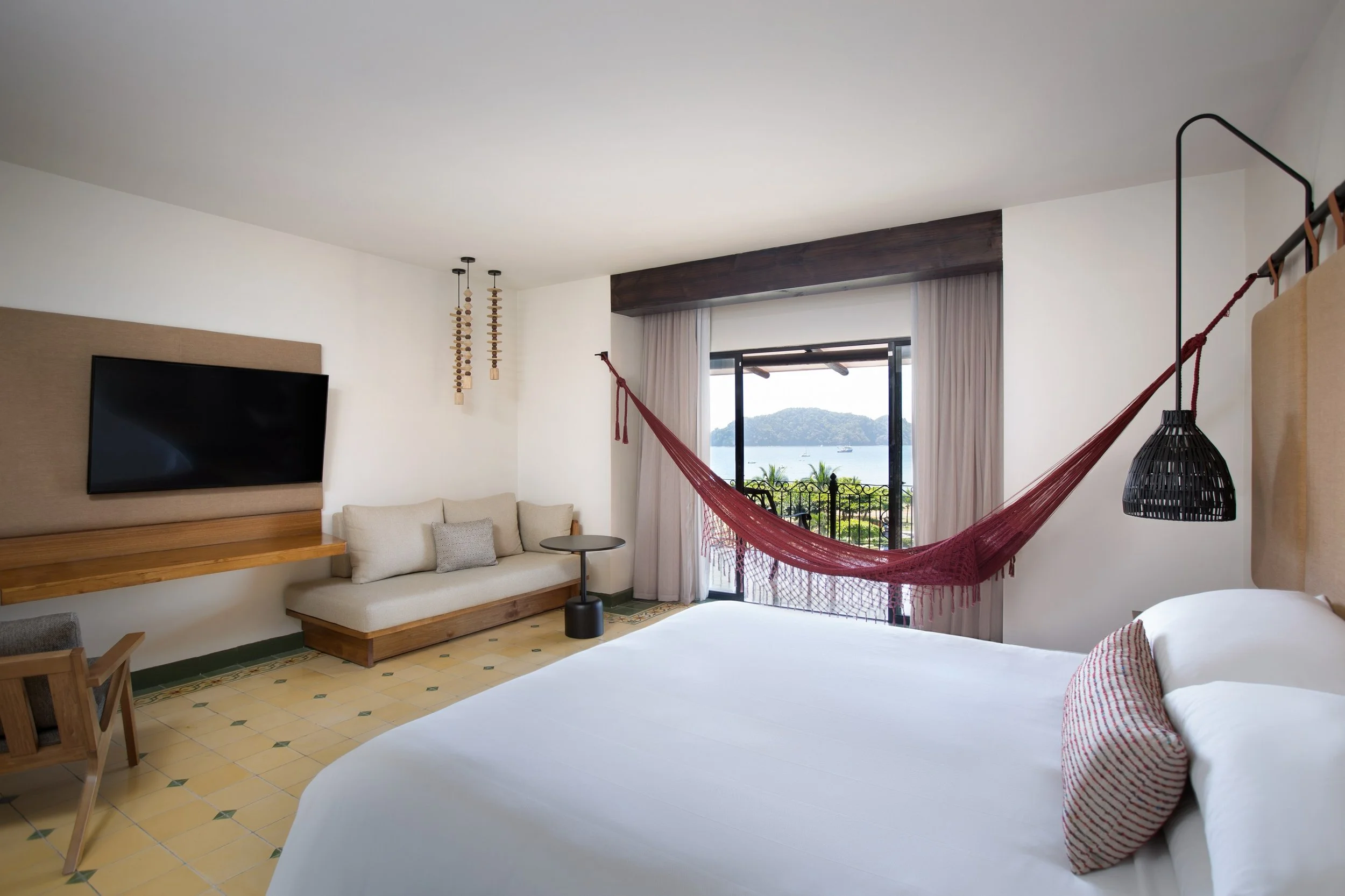 Hotel room with a large bed, hammock near balcony overlooking water, flat-screen TV, beige sofa, and modern decor.