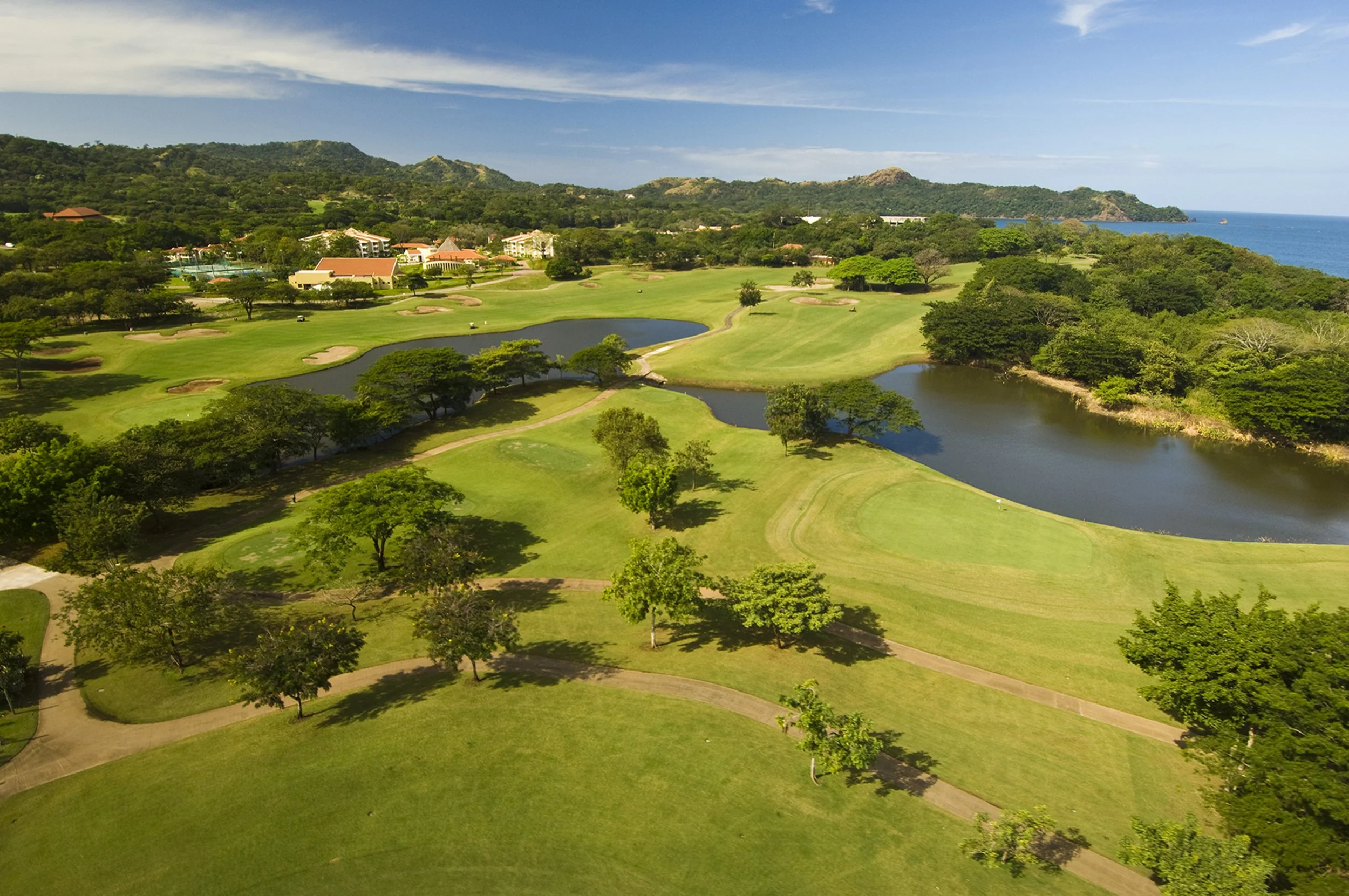Aerial view of a golf course with green fairways, water hazards, trees, and a distant coastline with hills and blue sky.