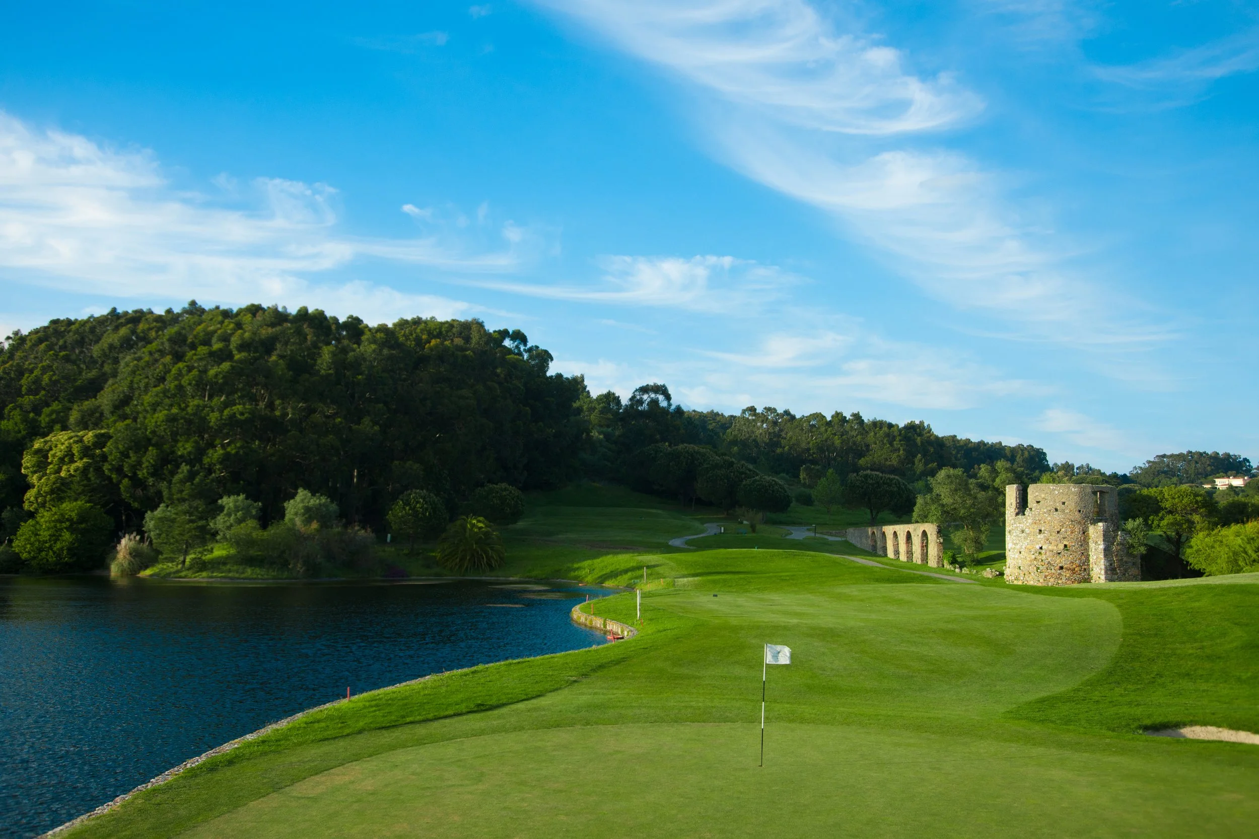 Scenic view of a golf course next to a river with green grass, a historic stone tower, and a background of lush trees and a blue sky with wispy clouds.
