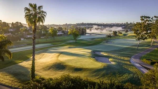 Scenic view of a golf course with lush green fairways, sand traps, a pond, and tall palm trees under a clear sky.