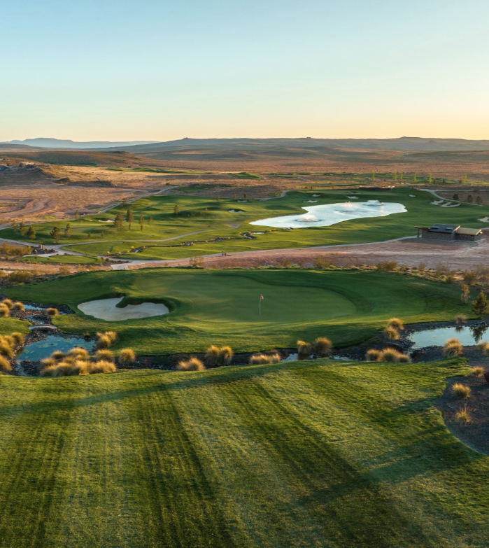 A scenic golf course with green fairways, sand traps, water hazards, and a club house in the distance, surrounded by desert landscape.